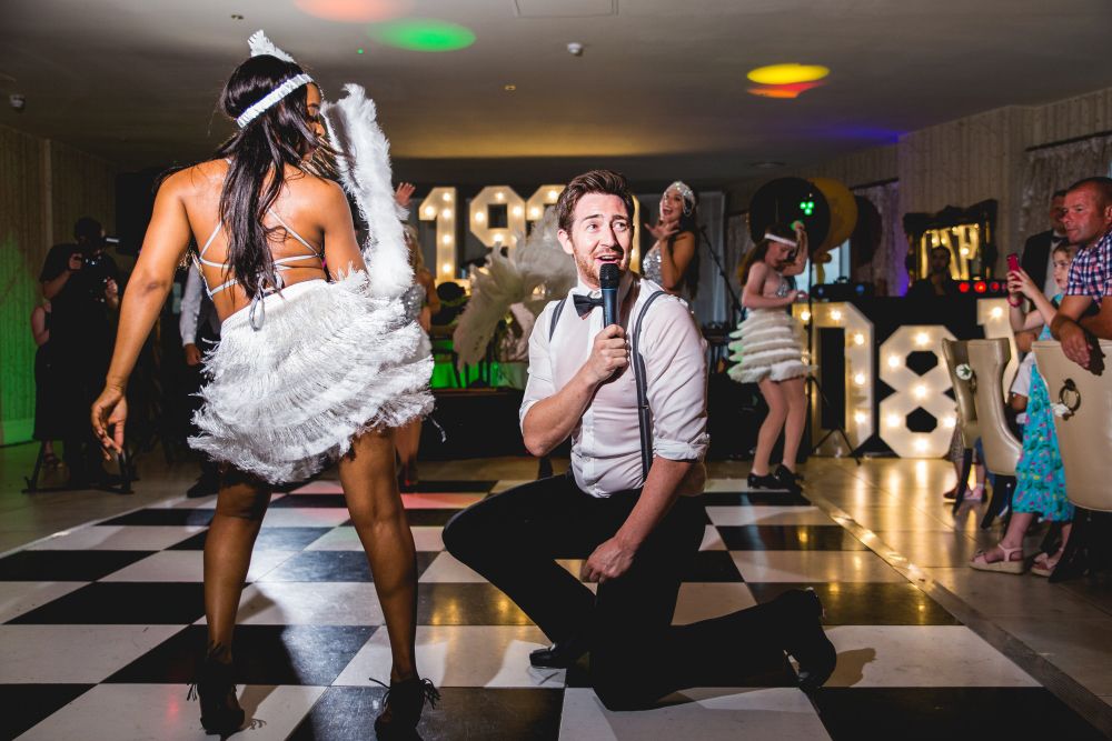A man kneels on a black and white checkered dance floor holding a microphone, looking up at a woman performing in a white feathered costume and headband, with other similarly dressed performers and large illuminated "18" numbers visible in the background. Image by Documentary Wedding Photographer Lewis Bishop Photography.
