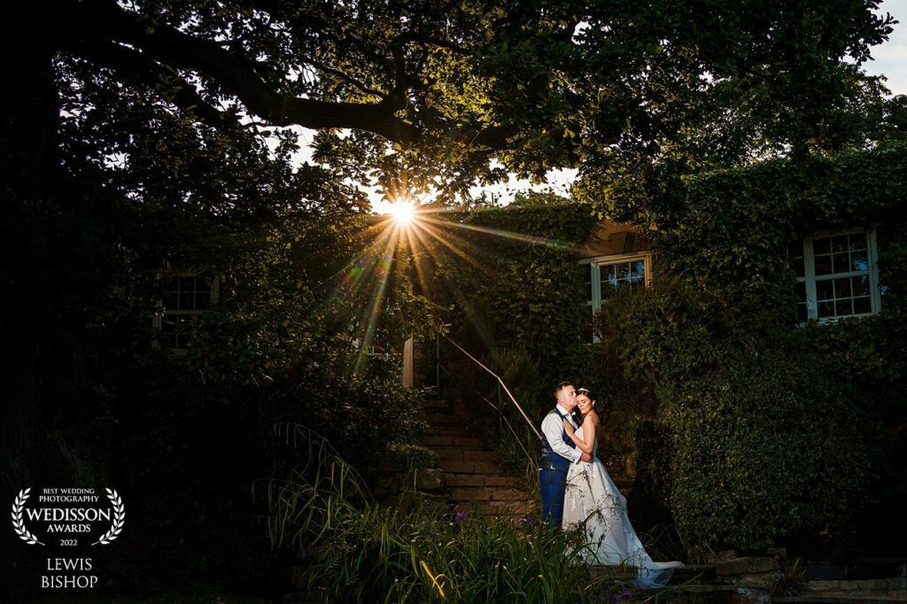 A newly married couple embraces under a large tree as the sun sets behind them, casting a warm glow. The groom wears suspenders while the bride is in a flowing white gown. They stand on stone steps surrounded by lush greenery. Award marks are in the lower left. Image by Documentary Wedding Photographer Lewis Bishop Photography.