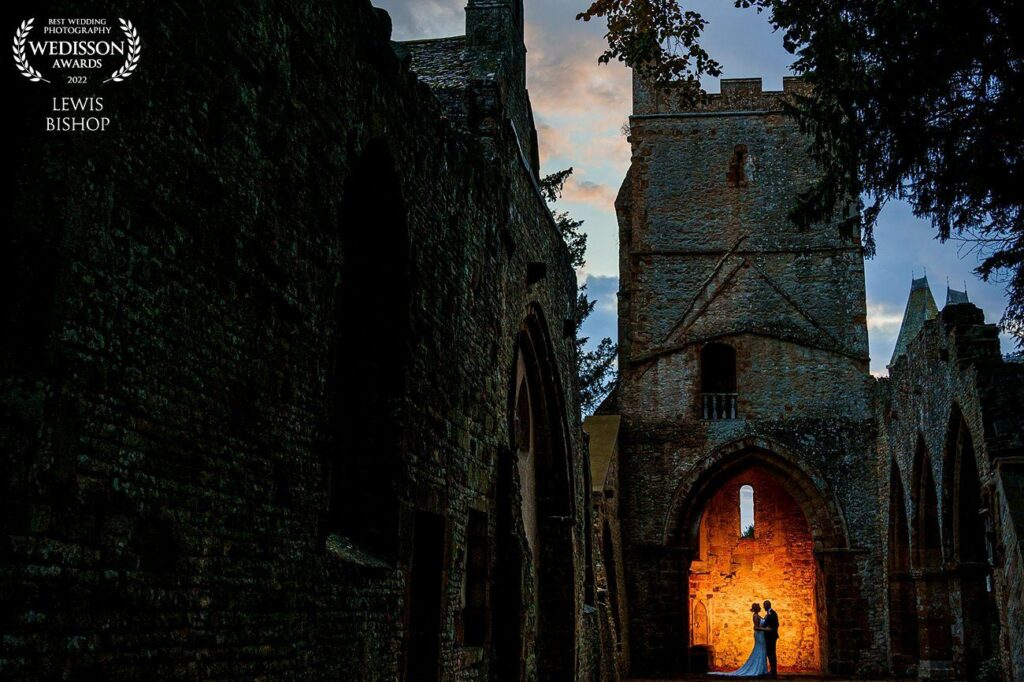 A couple dances in the ruins of an old stone building at dusk, illuminated by a warm golden light from an archway behind them. The dark structure contrasts with the twilight sky, creating a dramatic and romantic atmosphere. Image by Documentary Wedding Photographer Lewis Bishop Photography.