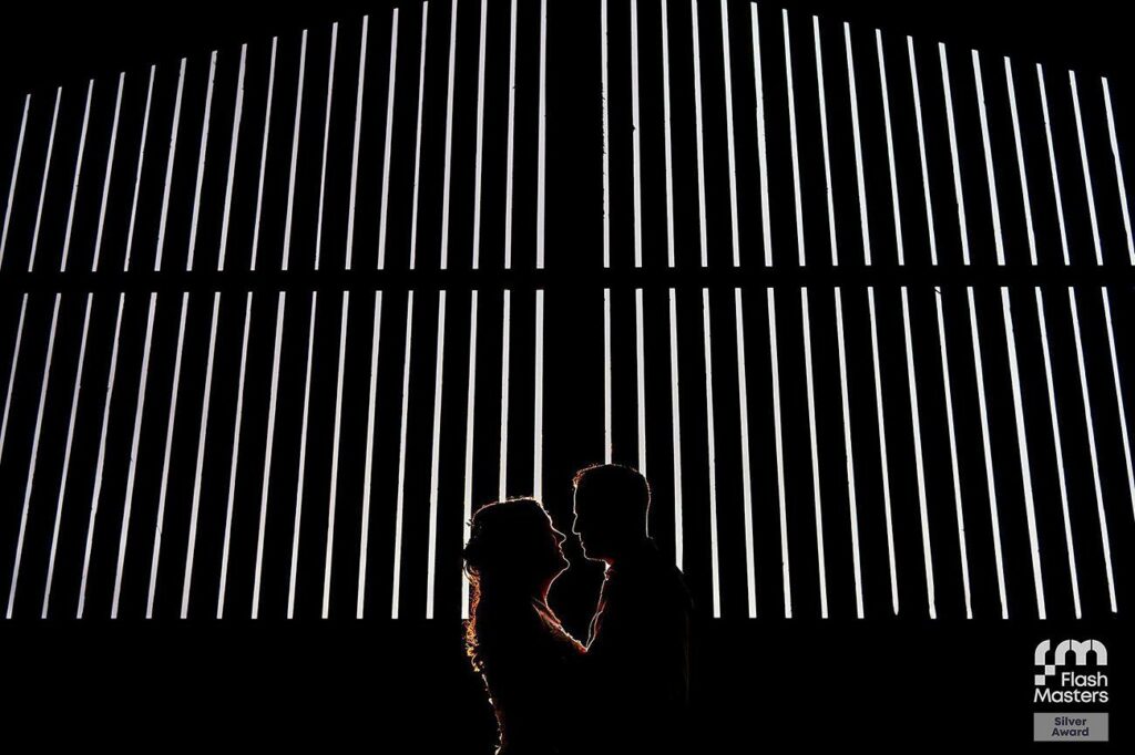 A silhouetted couple faces each other in front of vertical lines of bright white light in a dark setting. The man's outline is slightly more prominent than the woman's. The image features a "Flash Masters Silver Award" logo in the bottom right corner. Image by Documentary Wedding Photographer Lewis Bishop Photography.