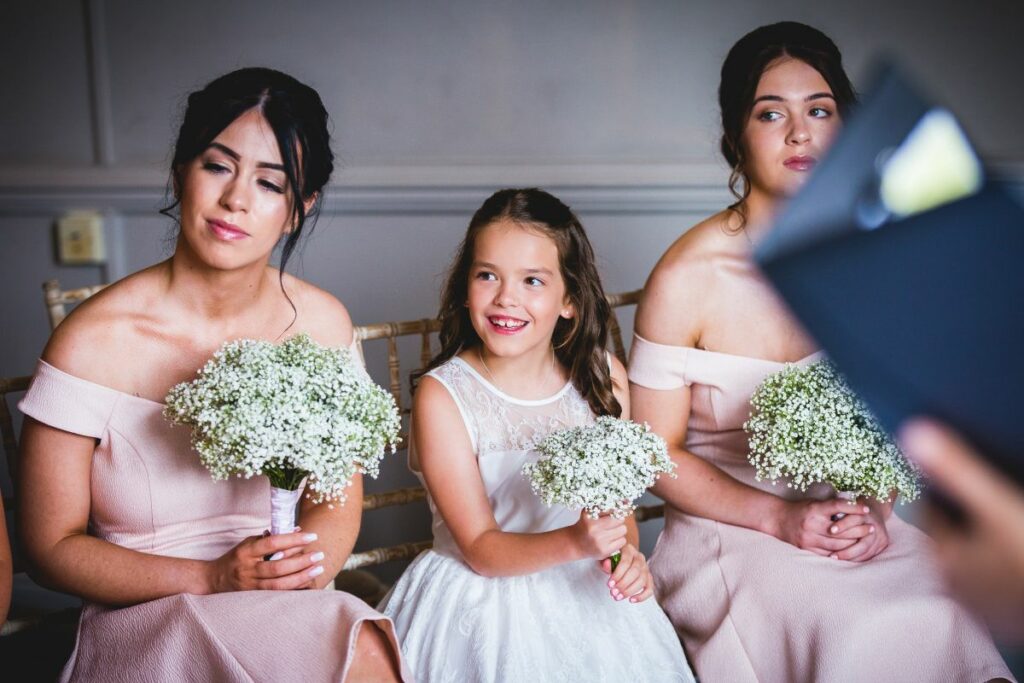 A young girl in a white dress is seated between two women in light pink dresses, all holding bouquets of baby's breath flowers. The girl is smiling while the women look off to the side. They appear to be part of a formal event, possibly a wedding. Image by Documentary Wedding Photographer Lewis Bishop Photography.