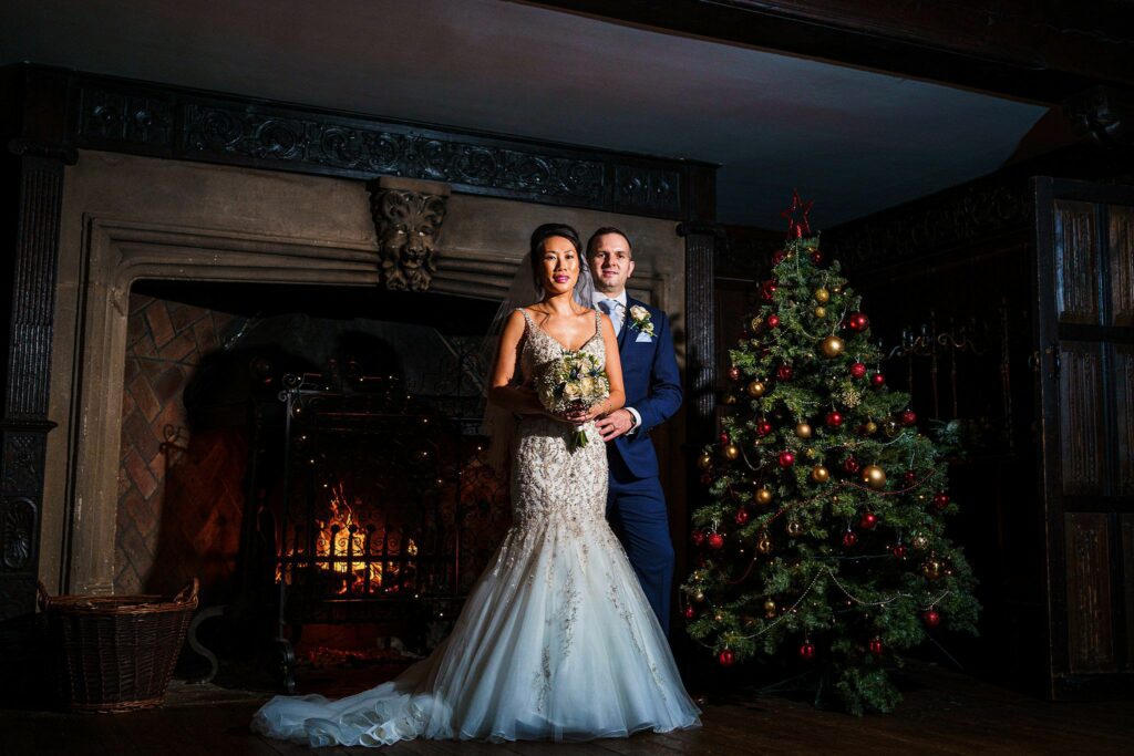 A bride and groom stand together in front of a lit fireplace in a dark wood-paneled room. The bride wears a white wedding dress and holds a bouquet, while the groom is in a blue suit. A decorated Christmas tree stands to their right, adding a festive touch. Image by Documentary Wedding Photographer Lewis Bishop Photography.