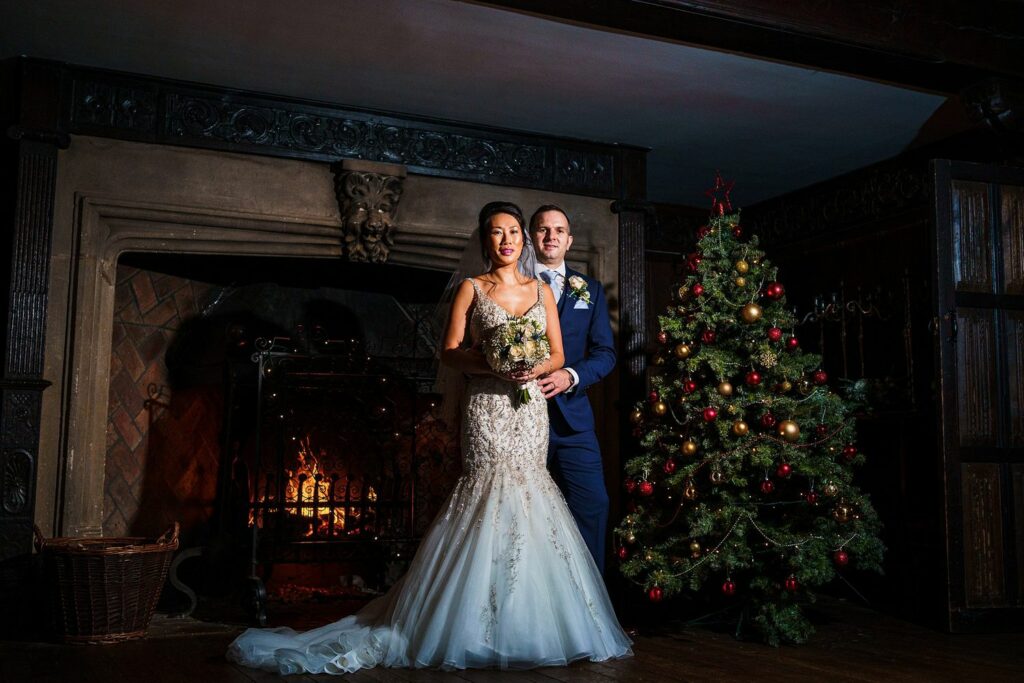 A bride in a white gown and a groom in a dark suit stand in front of a lit fireplace, holding a bouquet, next to a decorated Christmas tree in a dimly lit room with dark wood paneling. Image by Documentary Wedding Photographer Lewis Bishop Photography.
