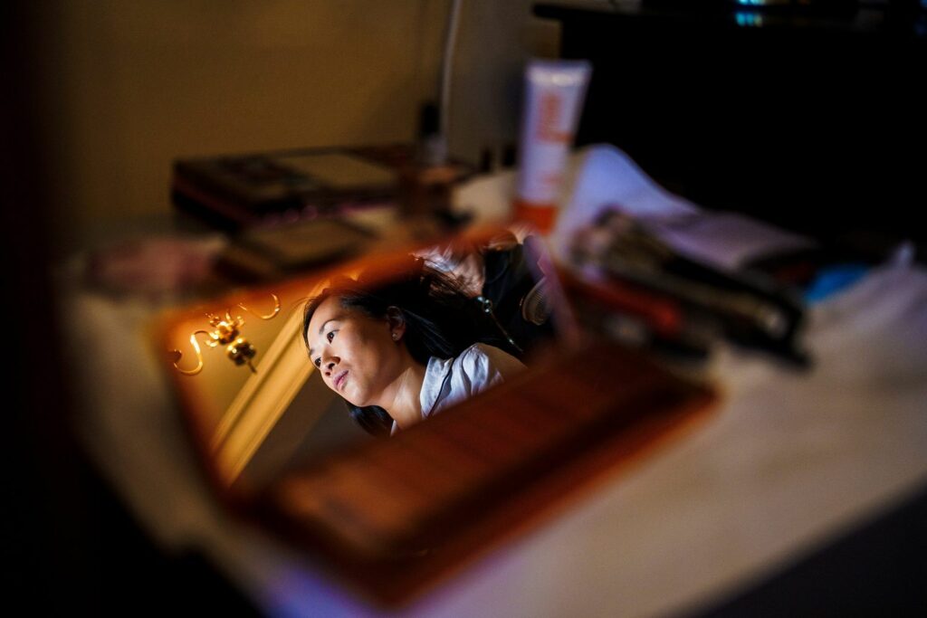 A young woman is reflected in a makeup mirror as she does her makeup. Various makeup items, including brushes and palettes, are scattered on the table around the mirror. A warm, softly lit room with a chandelier is visible in the background. Image by Documentary Wedding Photographer Lewis Bishop Photography.