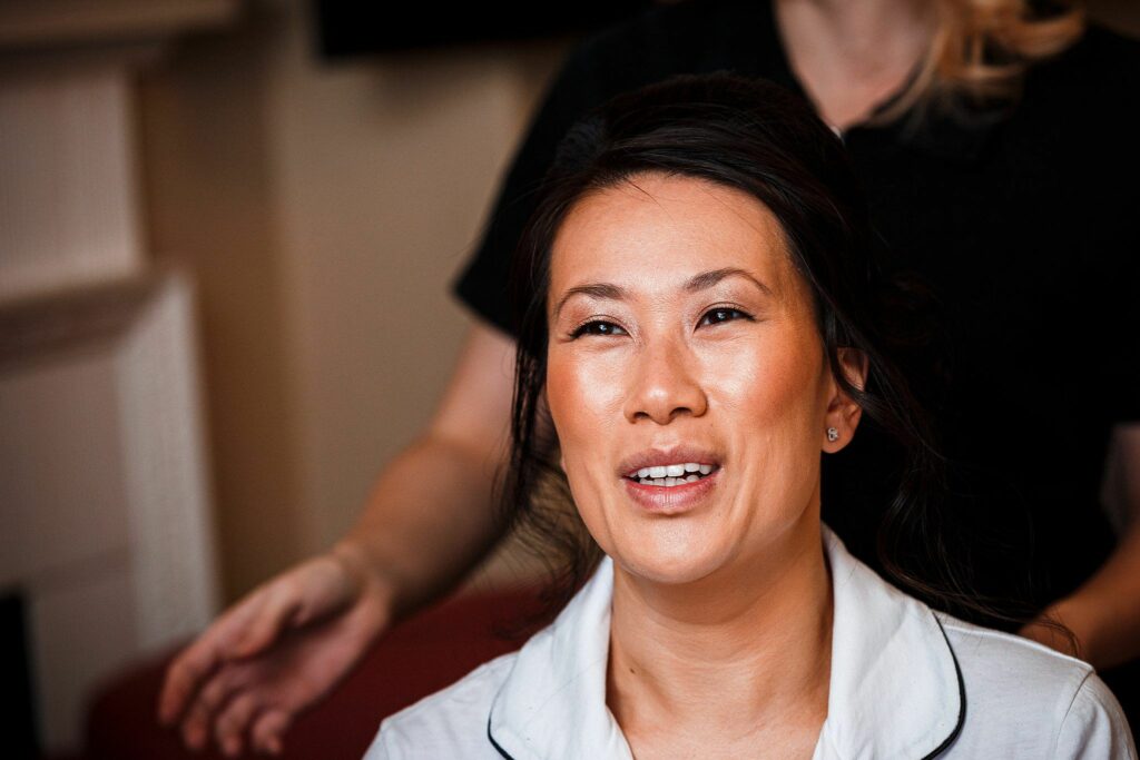 A woman with dark hair is smiling while someone behind her adjusts her hair. She is wearing a light-colored top with a dark trim and sitting in an indoor setting. The background is softly blurred, focusing attention on the woman. Image by Documentary Wedding Photographer Lewis Bishop Photography.