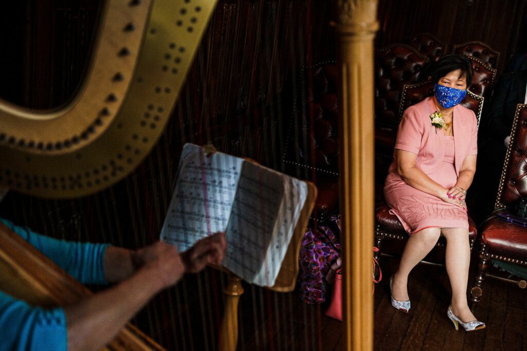 A woman in a pink dress and blue face mask sits on an ornate wooden chair, watching a harpist whose hands are visible playing the harp in the foreground. A sheet of music is mounted on the harp, and the scene is set in a richly decorated room. Image by Documentary Wedding Photographer Lewis Bishop Photography.