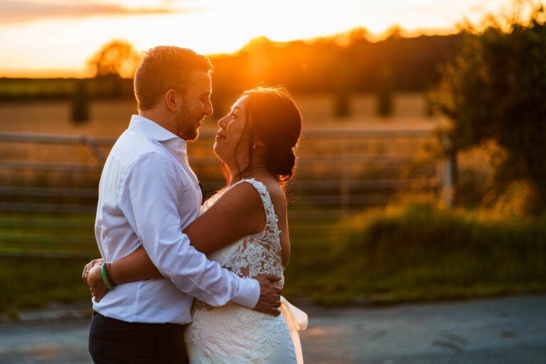A couple embraces outdoors at sunset, dressed in wedding attire, with a rural backdrop of a field and fence. Image by Documentary Wedding Photographer Lewis Bishop Photography.