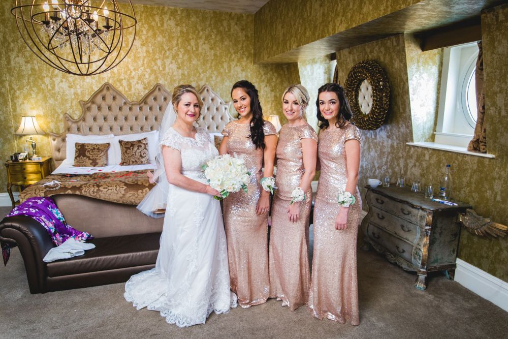 A bride in a white dress stands beside three bridesmaids in matching champagne-colored sequin dresses. They are standing in an elegant room with vintage decor, including a tufted headboard, chandelier, and ornate mirror. The bridesmaids wear corsages on their wrists. Image by Documentary Wedding Photographer Lewis Bishop Photography.