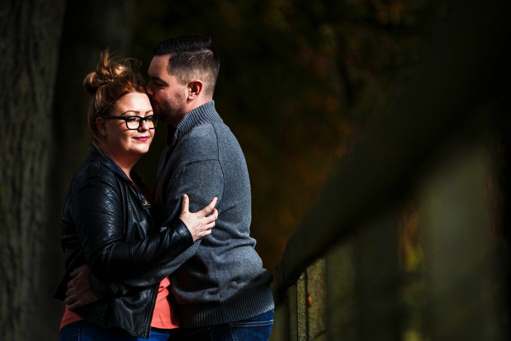 A couple embraces outdoors near a wooded area. The woman, wearing glasses and a black jacket, hugs the man, who is in a grey sweater. The man leans in to kiss the woman's forehead. They both appear content and happy in each other's company. Image by Documentary Wedding Photographer Lewis Bishop Photography.