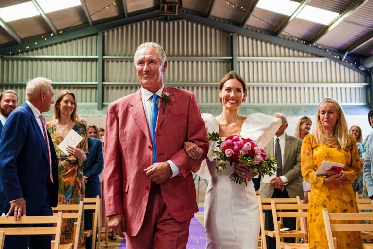A bride in a white dress, holding a bouquet of pink flowers, walks down an aisle arm-in-arm with an older man in a red suit. They are smiling, and guests in colorful attire are seated and standing, watching them in a well-lit indoor venue. Image by Documentary Wedding Photographer Lewis Bishop Photography.
