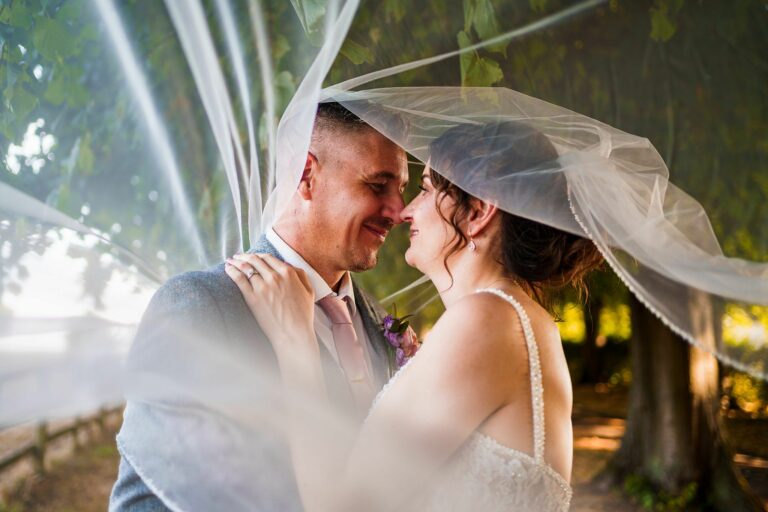 A bride and groom share a close, joyful moment under the bride's veil in a forested area. The couple appears happy and intimate, with soft light filtering through the trees behind them. The greenery and soft veil create a romantic ambiance. Image by Documentary Wedding Photographer Lewis Bishop Photography.