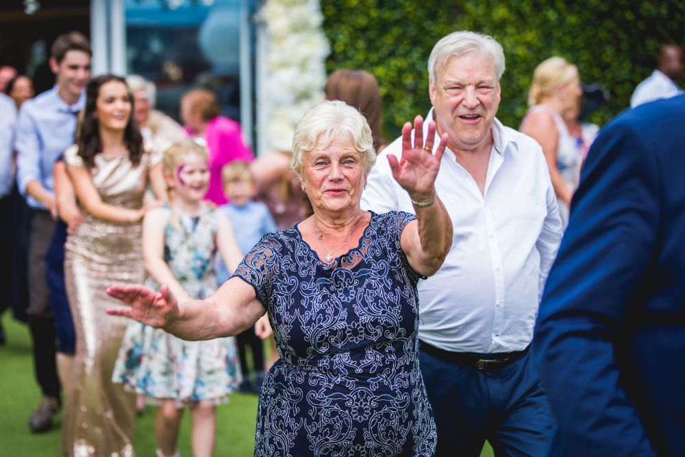 An elderly woman and man, both smiling and waving, lead a line of dancing people outdoors. The woman is wearing a patterned dress, and the man is in a white shirt. Other adults and children in festive attire follow them, enjoying the celebration. Image by Documentary Wedding Photographer Lewis Bishop Photography.