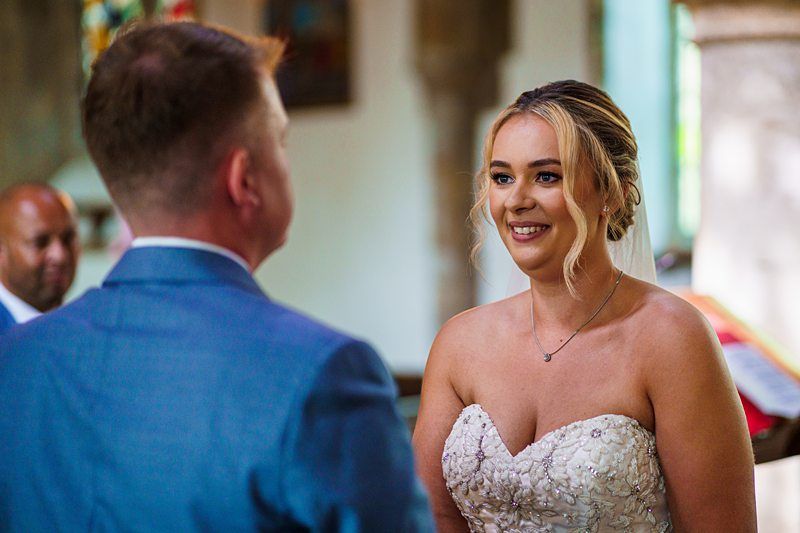 A bride in a strapless white gown smiles at her groom, who is dressed in a blue suit, during their wedding ceremony. The setting appears to be indoors, decorated with colorful elements, and a seated guest is visible in the background. Image by Documentary Wedding Photographer Lewis Bishop Photography.