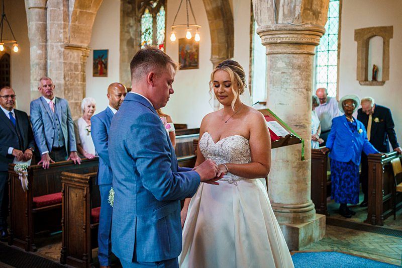A bride and groom stand at the altar in a church, exchanging rings. The groom is in a blue suit, while the bride wears a strapless white gown. Guests, including older adults, are seated in wooden pews, watching. The church interior features stone columns and arched windows. Image by Documentary Wedding Photographer Lewis Bishop Photography.