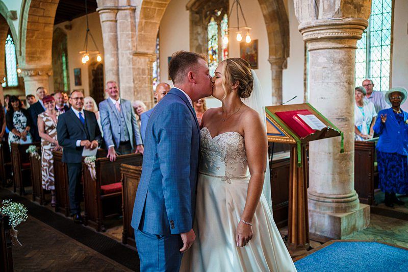 A bride and groom share a kiss during their wedding ceremony inside a church. The bride is wearing a strapless white dress and the groom is in a blue suit. Guests seated in pews smile and watch them. Stained glass windows and stone columns are visible in the background. Image by Documentary Wedding Photographer Lewis Bishop Photography.