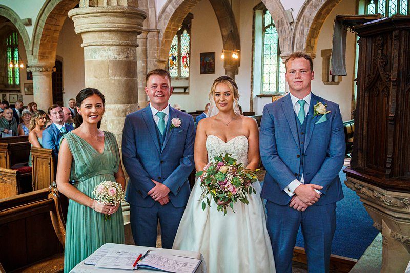 A newlywed couple and their two attendants stand inside a church. The bride is in a white gown holding a bouquet, and the groom is in a blue suit with a pink boutonniere. The bridesmaid, in a green dress, holds a bouquet, and the groomsman is in a blue suit. Image by Documentary Wedding Photographer Lewis Bishop Photography.