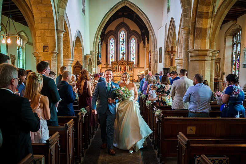 A happy couple walks down the aisle together in a beautifully decorated church, surrounded by smiling guests. The groom is wearing a suit and boutonniere, while the bride is in a white wedding gown holding a bouquet. Stained glass windows are visible in the background. Image by Documentary Wedding Photographer Lewis Bishop Photography.