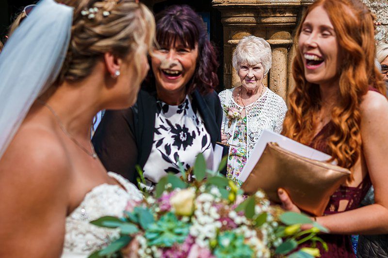 A bride in a white wedding dress and veil is holding a bouquet and smiling, facing two women who are laughing. An older woman with white hair, wearing a white cardigan, is standing in the background, observing the scene. Image by Documentary Wedding Photographer Lewis Bishop Photography.
