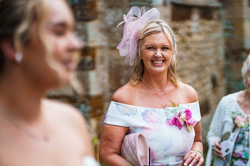 A woman wearing a floral off-the-shoulder dress and a pink fascinator smiles while holding a pink clutch bag. She stands outdoors with a blurred background, and another person is partially visible in the foreground. Image by Documentary Wedding Photographer Lewis Bishop Photography.