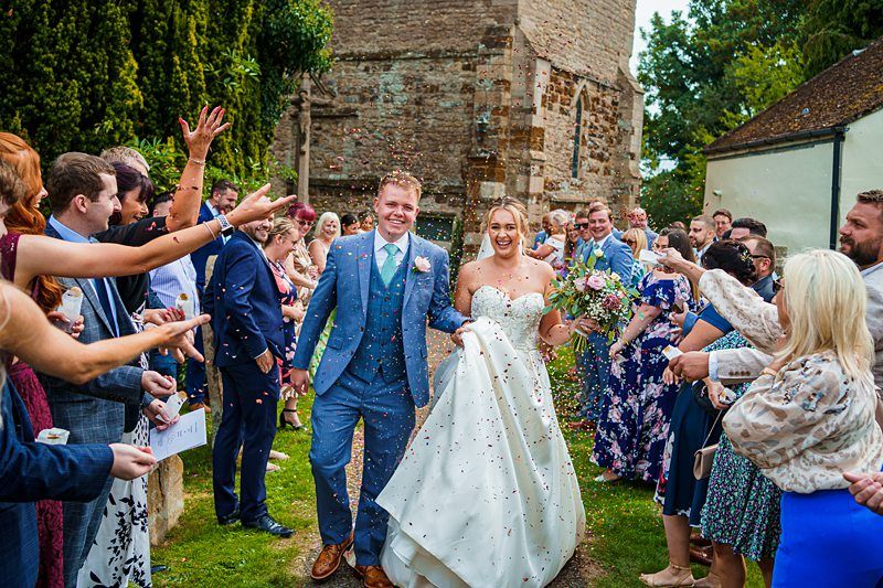 A newlywed couple, dressed in a blue suit and white wedding gown, joyfully walks down a path as guests on both sides shower them with confetti. Smiling guests, holding cameras and phones, celebrate outside a rustic stone building and lush greenery. Image by Documentary Wedding Photographer Lewis Bishop Photography.