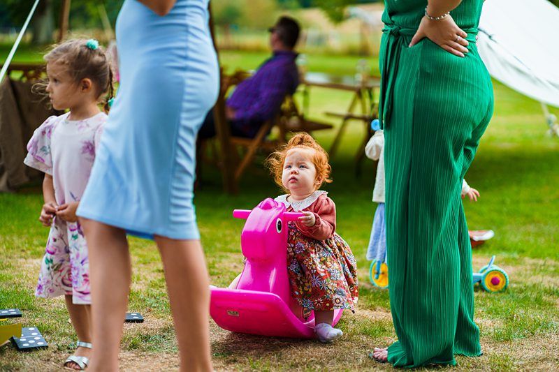 A small child with red hair sits on a pink toy horse in a grassy outdoor area. The child is looking up, surrounded by adults and other children. The scene is colorful, with adults in blue and green outfits and other children nearby. Image by Documentary Wedding Photographer Lewis Bishop Photography.