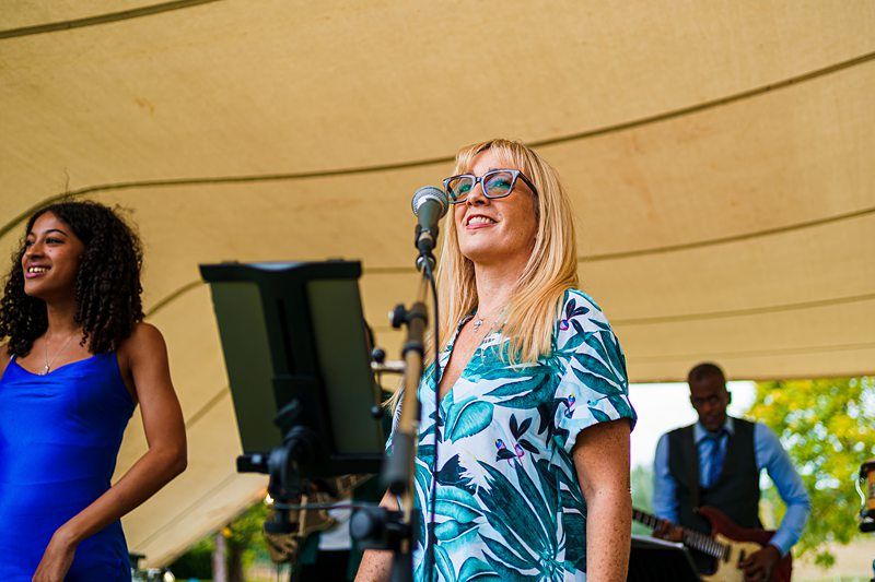 Two women, one in a blue dress and the other in a tropical print dress, sing into microphones under a tent. A man in the background plays an electric guitar. The setting appears to be an outdoor event. Image by Documentary Wedding Photographer Lewis Bishop Photography.
