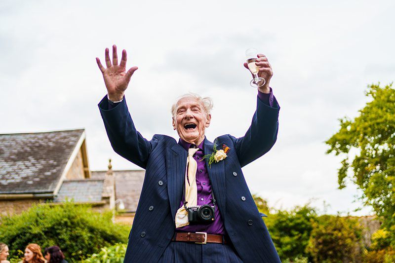 An elderly man joyfully raises his hands in the air, one arm waving and the other holding a glass. He is dressed in a suit with a flower boutonnière and has a camera around his neck. The scene appears to be outdoors, with greenery and buildings in the background. Image by Documentary Wedding Photographer Lewis Bishop Photography.