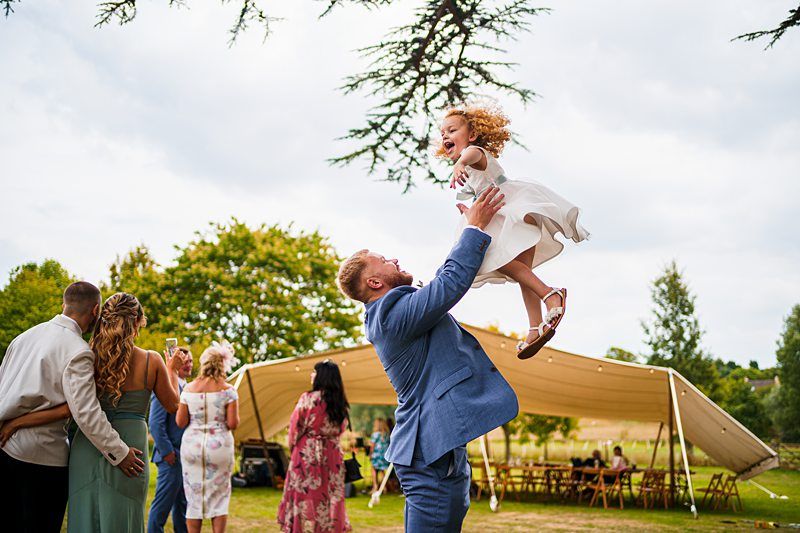 A man in a blue suit joyfully lifts a laughing young girl in a white dress above his head. They are outdoors at a gathering or event, with a large tent, trees, and guests dancing and mingling in the background. The atmosphere is lively and festive. Image by Documentary Wedding Photographer Lewis Bishop Photography.