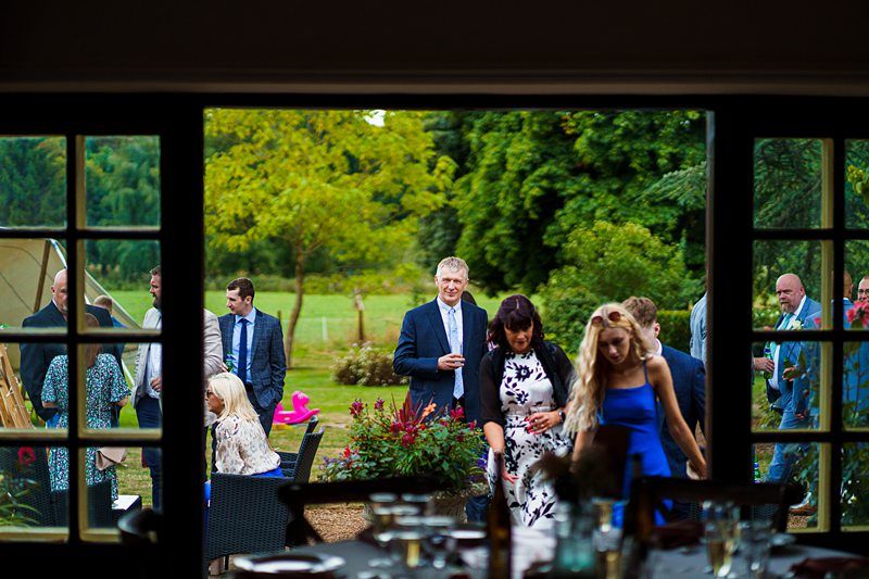 A group of people gather outdoors at a garden party. Visible through a large window, some are standing and chatting while others are seated. Trees and greenery form the background, with individuals dressed in formal and semi-formal attire. Image by Documentary Wedding Photographer Lewis Bishop Photography.