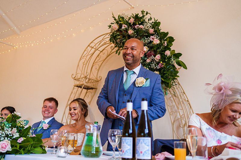 A smiling man in a blue suit and green tie stands to give a speech at a wedding reception. Behind him is a floral arch. Seated at the table are the bride in a white dress, another man, and two women, one of whom is wearing a fancy hat. Image by Documentary Wedding Photographer Lewis Bishop Photography.