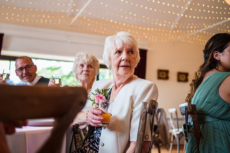 An elderly woman with white hair, wearing a white jacket, holds a drink while seated at a table during a social event. She is surrounded by other guests in a warmly lit room decorated with string lights on the ceiling. Image by Documentary Wedding Photographer Lewis Bishop Photography.