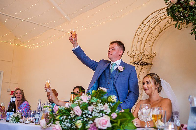 A groom in a blue suit raises a toast with a champagne glass at a wedding reception. The bride, dressed in a white gown and veil, sits beside him, smiling. Other guests, seated at a decorated table with flowers and drinks, join in the celebration. String lights adorn the ceiling. Image by Documentary Wedding Photographer Lewis Bishop Photography.