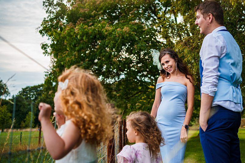 A woman in a light blue dress stands next to a man in a blue shirt and vest, smiling and interacting with three young children. The scene is set outdoors on a sunny day, with trees and a grassy area in the background. Image by Documentary Wedding Photographer Lewis Bishop Photography.
