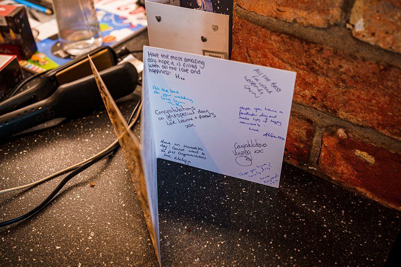 A greeting card stands open on a countertop beside a brick wall. The card contains handwritten messages, well-wishes, and signatures. Nearby, there are various other items including a stapler, a plastic bottle, and other stationery. Image by Documentary Wedding Photographer Lewis Bishop Photography.