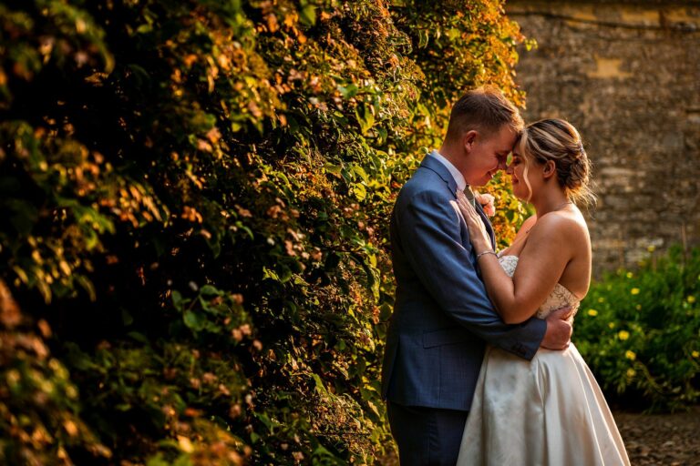 A couple embraces tenderly in front of sunlit ivy-covered walls. The man in a gray suit and the woman in a white wedding dress lean their foreheads together, sharing an intimate moment as warm, golden light bathes the scene. Image by Documentary Wedding Photographer Lewis Bishop Photography.