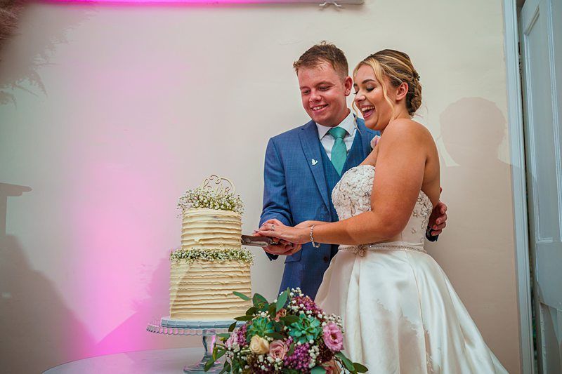 A newlywed couple is cutting their wedding cake together. The bride is wearing a white strapless gown and is smiling joyfully, while the groom, dressed in a blue suit and green tie, stands beside her, smiling. The two-tiered cake is decorated with flowers. Image by Documentary Wedding Photographer Lewis Bishop Photography.