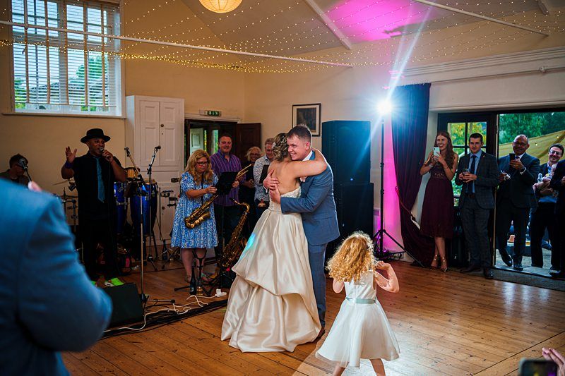A bride and groom share a joyful embrace while dancing at their wedding reception. The bride wears a white gown, and the groom is in a gray suit. A young girl dances nearby. Guests and musicians with instruments applaud and celebrate in the background. Image by Documentary Wedding Photographer Lewis Bishop Photography.