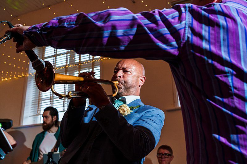A trumpet player wearing a blue suit and tie passionately performs with a brass trumpet. Another person in a purple striped shirt is in the foreground holding a microphone towards the trumpet. String lights decorate the background. Image by Documentary Wedding Photographer Lewis Bishop Photography.