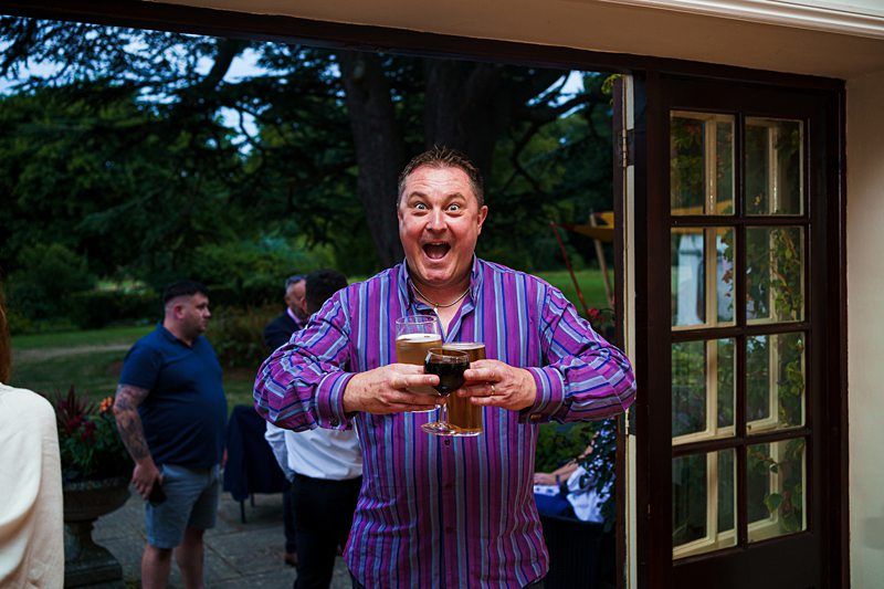 A man with a joyful expression stands indoors by an open door, holding three drinks and wearing a vibrant striped shirt. In the background, people are gathered outside in a garden setting, with greenery and trees visible. The atmosphere appears lively and festive. Image by Documentary Wedding Photographer Lewis Bishop Photography.