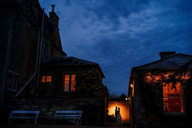 Silhouetted couple embracing under a warm light between two dark, stone buildings at night, with a deep blue sky overhead. White benches sit in the foreground, and windows in the buildings emit a soft glow. Image by Documentary Wedding Photographer Lewis Bishop Photography.