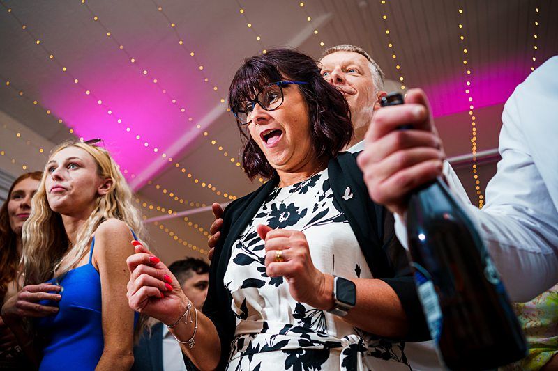 A group of people stand close together under string lights. A woman in the center, wearing glasses and a black-and-white dress, has a surprised expression. Beside her, a man holds a bottle, and others around them are smiling and looking in various directions. Image by Documentary Wedding Photographer Lewis Bishop Photography.