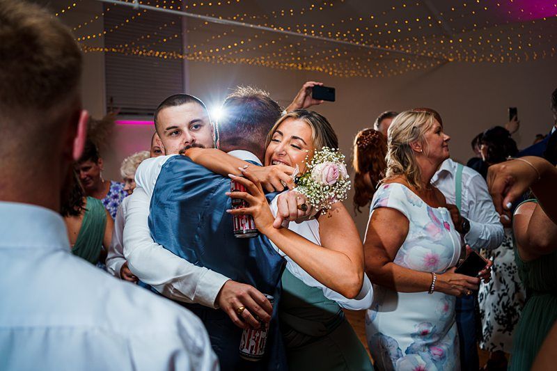 A group of people at a wedding reception are celebrating. A man in a blue suit is hugging a woman holding a small bouquet of flowers. Partygoers around them are dancing and talking under decorative string lights. Everyone appears joyful and festive. Image by Documentary Wedding Photographer Lewis Bishop Photography.