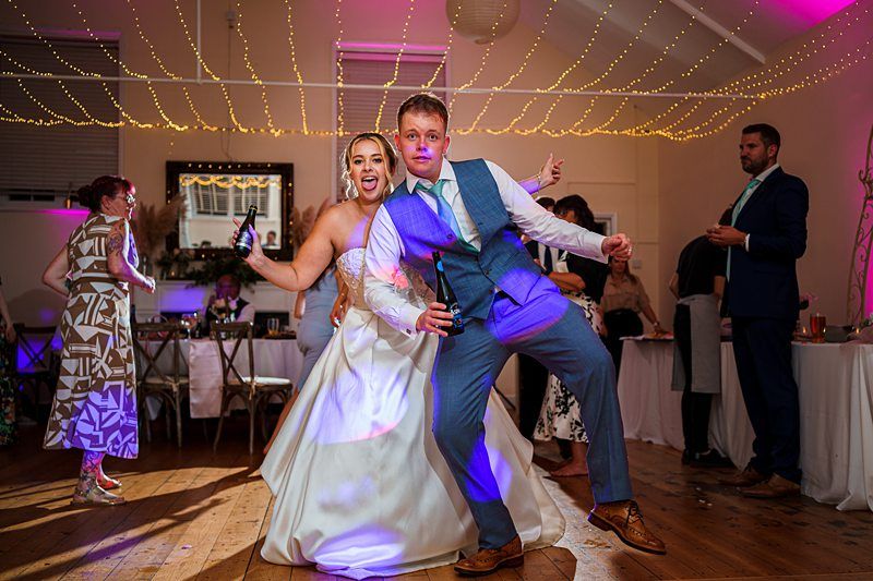 A bride and groom are joyfully dancing with drinks in their hands at a wedding reception. The bride is wearing a white dress and the groom is in a blue suit with a tie. Guests in elegant attire are in the background, beneath sparkling string lights. Image by Documentary Wedding Photographer Lewis Bishop Photography.