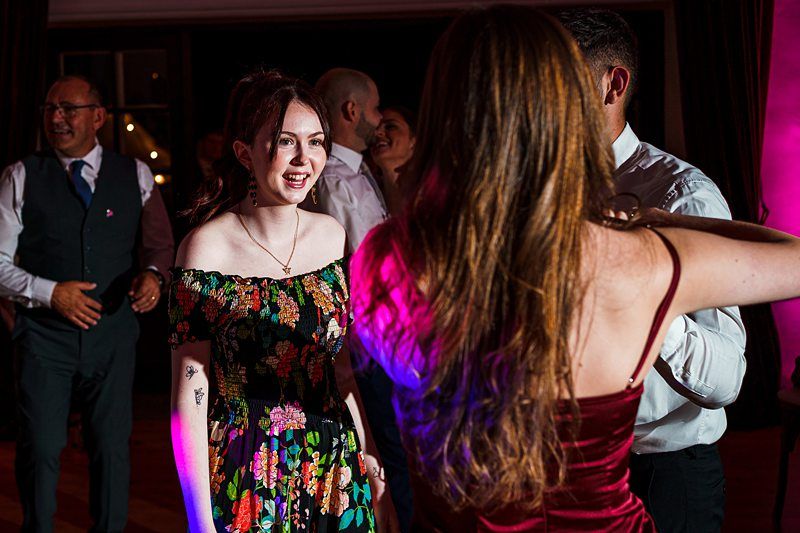 A woman in a floral dress smiles and talks to another woman wearing a red dress on a dance floor. Other people are dancing and chatting in the background, illuminated by colorful lights. Image by Documentary Wedding Photographer Lewis Bishop Photography.