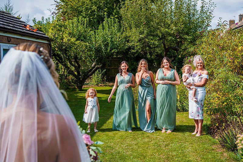 A bride in a white veil stands facing a group of bridesmaids and family in a lush garden. The bridesmaids wear matching green dresses, while an older woman in a floral dress holds a small child. A young girl in a white dress stands in front, looking towards the bride. Image by Documentary Wedding Photographer Lewis Bishop Photography.