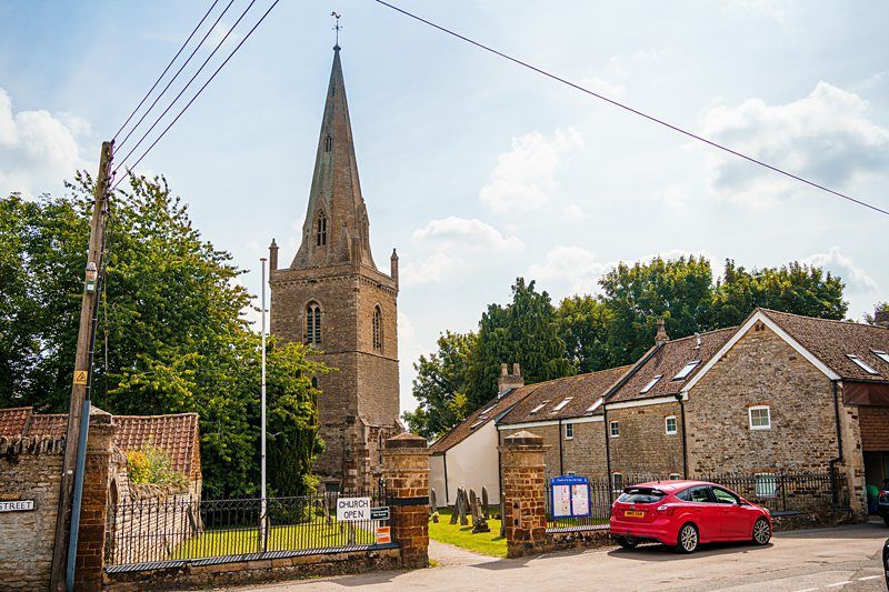 A village scene featuring a stone church with a tall steeple surrounded by greenery. In front, a sign reads "Church Open." Nearby, a red car is parked beside stone buildings with tiled roofs. Power lines cross the sky, and white clouds float above. Image by Documentary Wedding Photographer Lewis Bishop Photography.