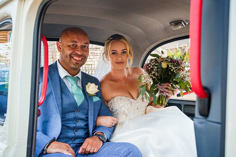 A smiling couple in wedding attire sit inside a car. The groom, wearing a blue suit with a green tie, has his arm around the bride, who is in a strapless white gown and holding a bouquet of flowers. Both look towards the camera, excited and happy. Image by Documentary Wedding Photographer Lewis Bishop Photography.