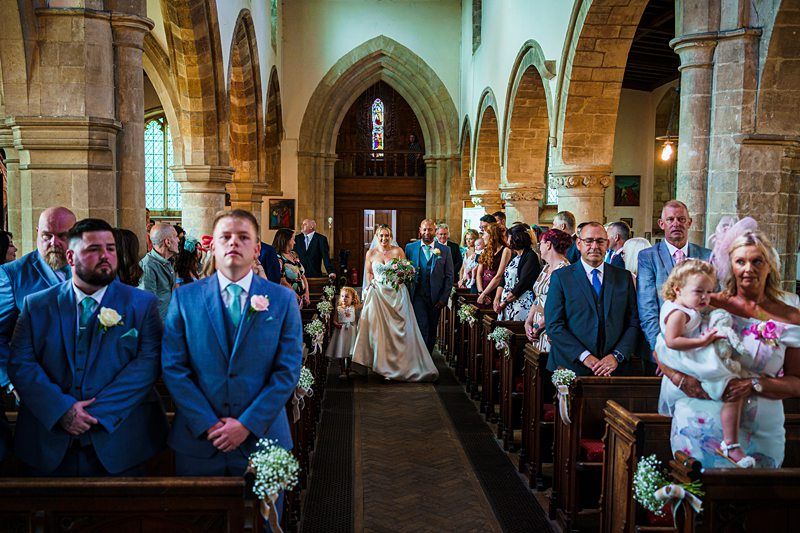 A bride walks down the aisle of a church with an older man, surrounded by standing guests. The groom, in a blue suit, waits at the altar with groomsmen. The church has arched doorways, stone columns, and small bouquets of white flowers attached to the pews. Image by Documentary Wedding Photographer Lewis Bishop Photography.