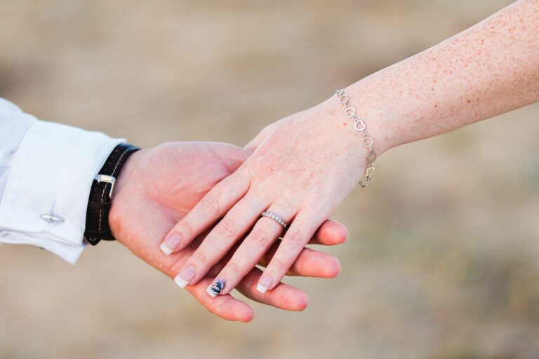 Close-up of two hands gently holding each other. One hand, adorned with a ring and a bracelet, is held by the other which has a watch on the wrist. The background is blurred, putting emphasis on the hands and jewelry. Image by Documentary Wedding Photographer Lewis Bishop Photography.