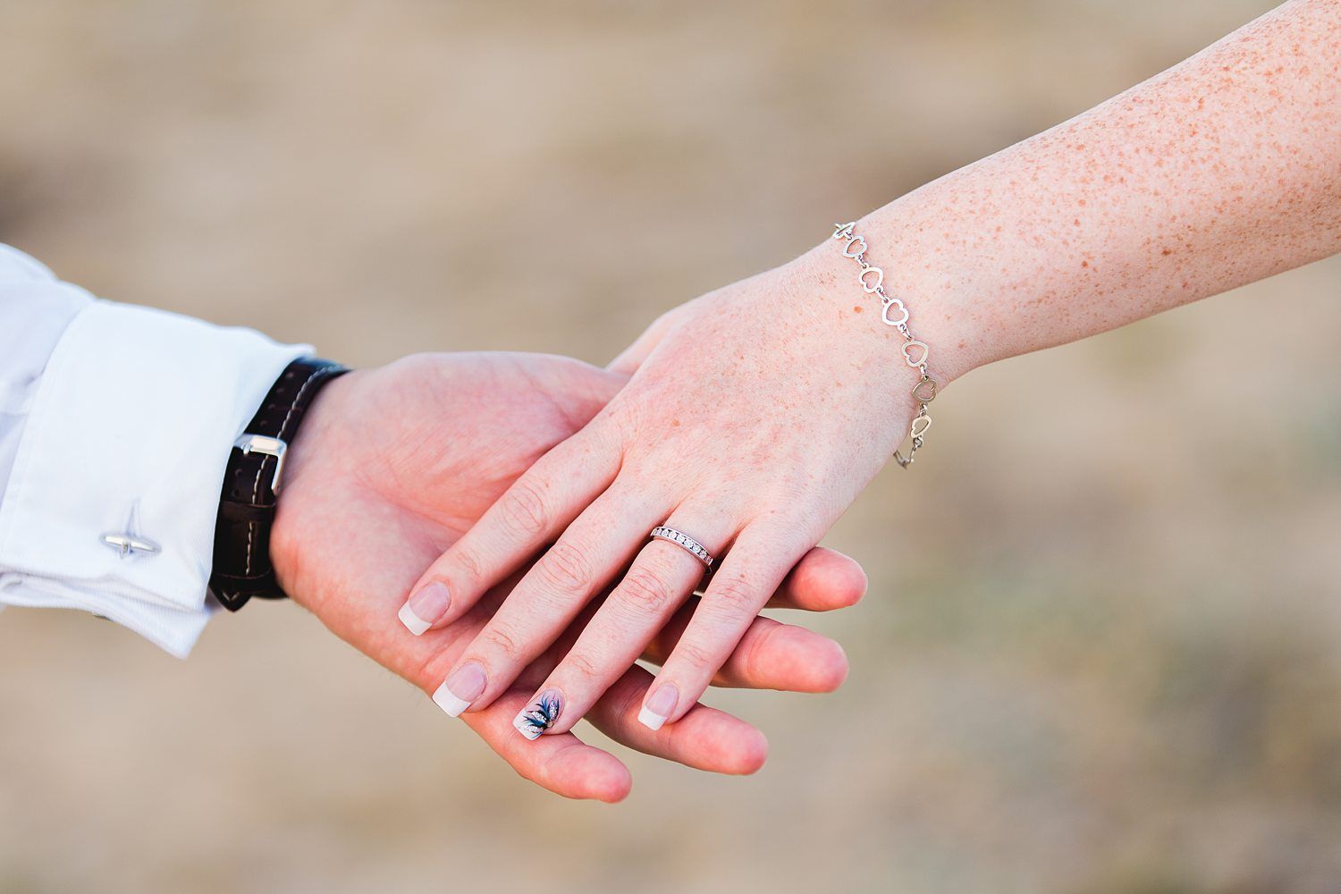 Close-up of two hands gently holding each other. One hand, adorned with a ring and a bracelet, is held by the other which has a watch on the wrist. The background is blurred, putting emphasis on the hands and jewelry. Image by Documentary Wedding Photographer Lewis Bishop Photography.
